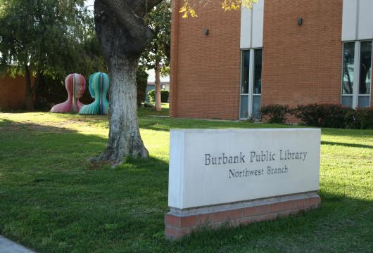 A sign in front of the Burbank Public Library Northwest Branch reads "Burbank Public Library Northwest Branch" with abstract colorful sculptures in the background.