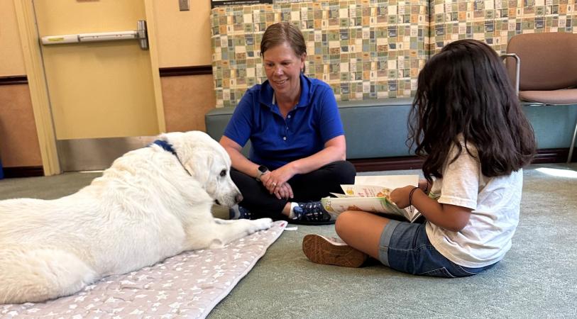 A white dog lies on a mat while a person in a blue shirt and a child sit on the floor reading a book.