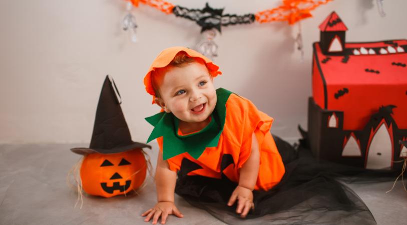 A smiling baby in an orange Halloween costume with a green collar and orange hat kneels on a black sheet, next to a carved jack-o’-lantern bucket and a red haunted-house decoration.