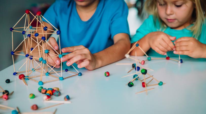 Two people sit at a table building colorful geometric structures from wooden sticks and beads, with hands moving small parts and finished frame models nearby.