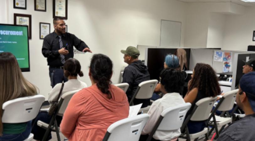A trainer stands in front of a classroom of seated attendees, gesturing while speaking, with chairs facing a whiteboard and projector screens on the walls.