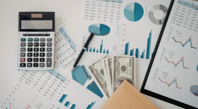 A desk with a calculator, spreadsheets and bar and line charts on screens, a calculator keypad, and a bundle of banknotes with coins.