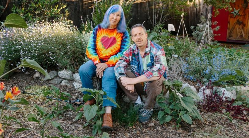Two people sitting among colorful garden plants and flowers, with a rustic wooden fence and a red decorative panel in the background.