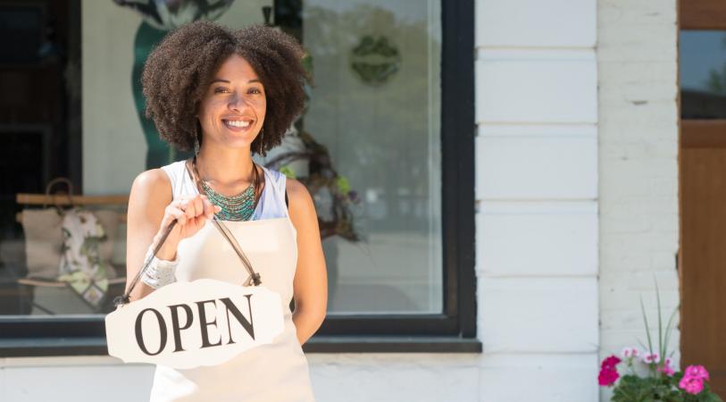 young lady wearing apron and holding an open sign in front of a storefront