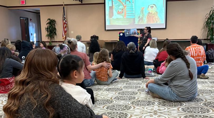 A group of people, including adults and children, are sitting on the floor watching a presenter at a "Family Storytime" event hosted by the Buena Vista Branch Library. A screen behind the presenter displays the event title and dates.