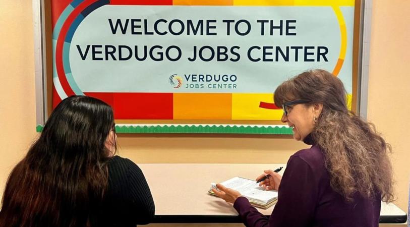 Two individuals sit at a table in front of a "Welcome to the Verdugo Jobs Center" sign. One person is writing on a notepad.