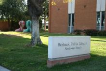 A sign in front of the Burbank Public Library Northwest Branch reads "Burbank Public Library Northwest Branch" with abstract colorful sculptures in the background.